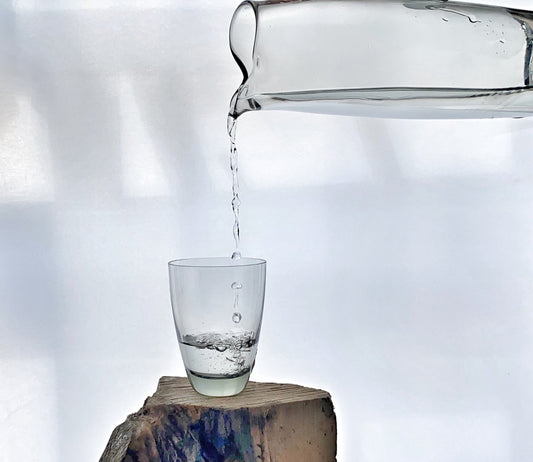 Clear glass carafe pouring water into a tumbler balanced on a natural wood block.