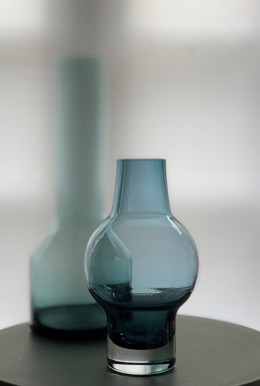 Blue glass vase in focus with a second vase by the same designer softly blurred in the background.